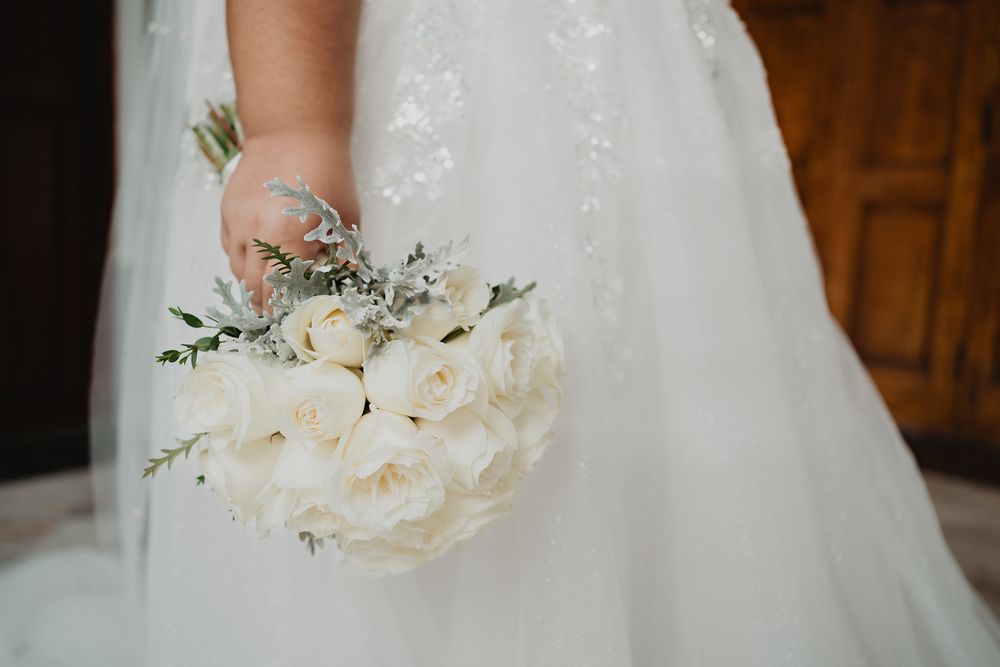 Close-up of a white wedding bouquet with roses and greenery held by person in flowing white dress.