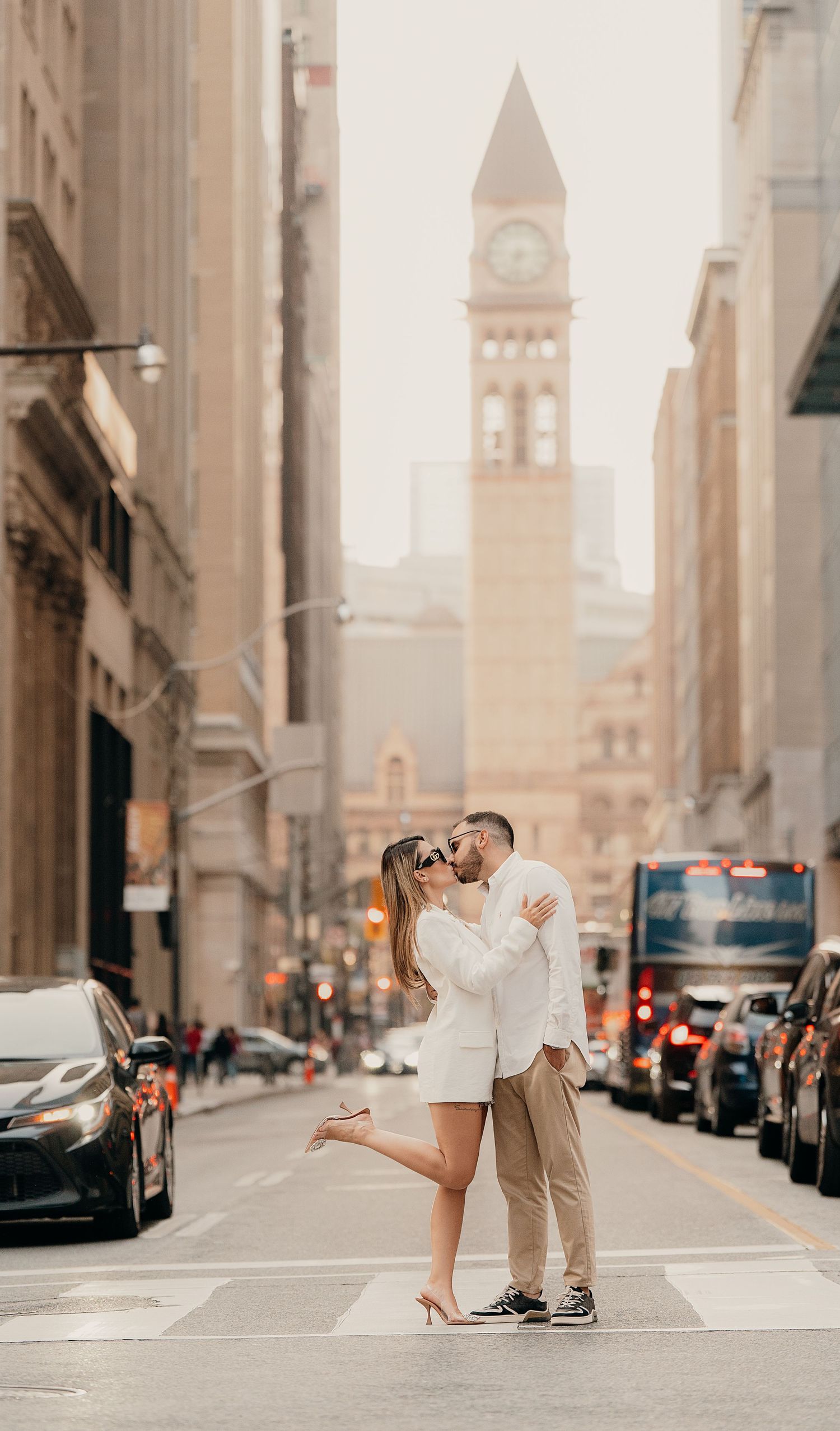 A couple poses romantically in the middle of a city street with tall buildings and a vintage clock tower in the background.