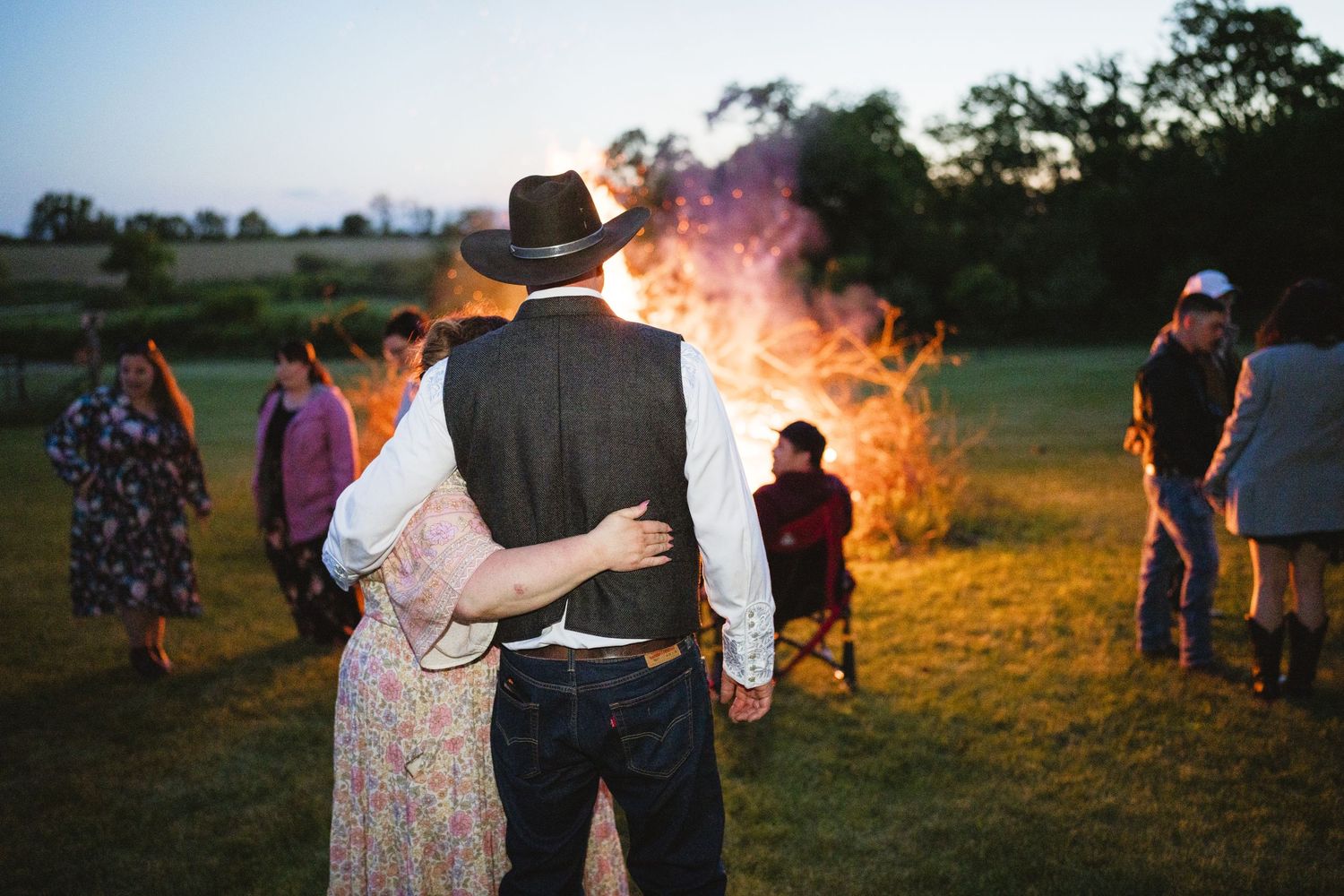 Two people stand together watching a large bonfire illuminate a rural field at golden sunset.
