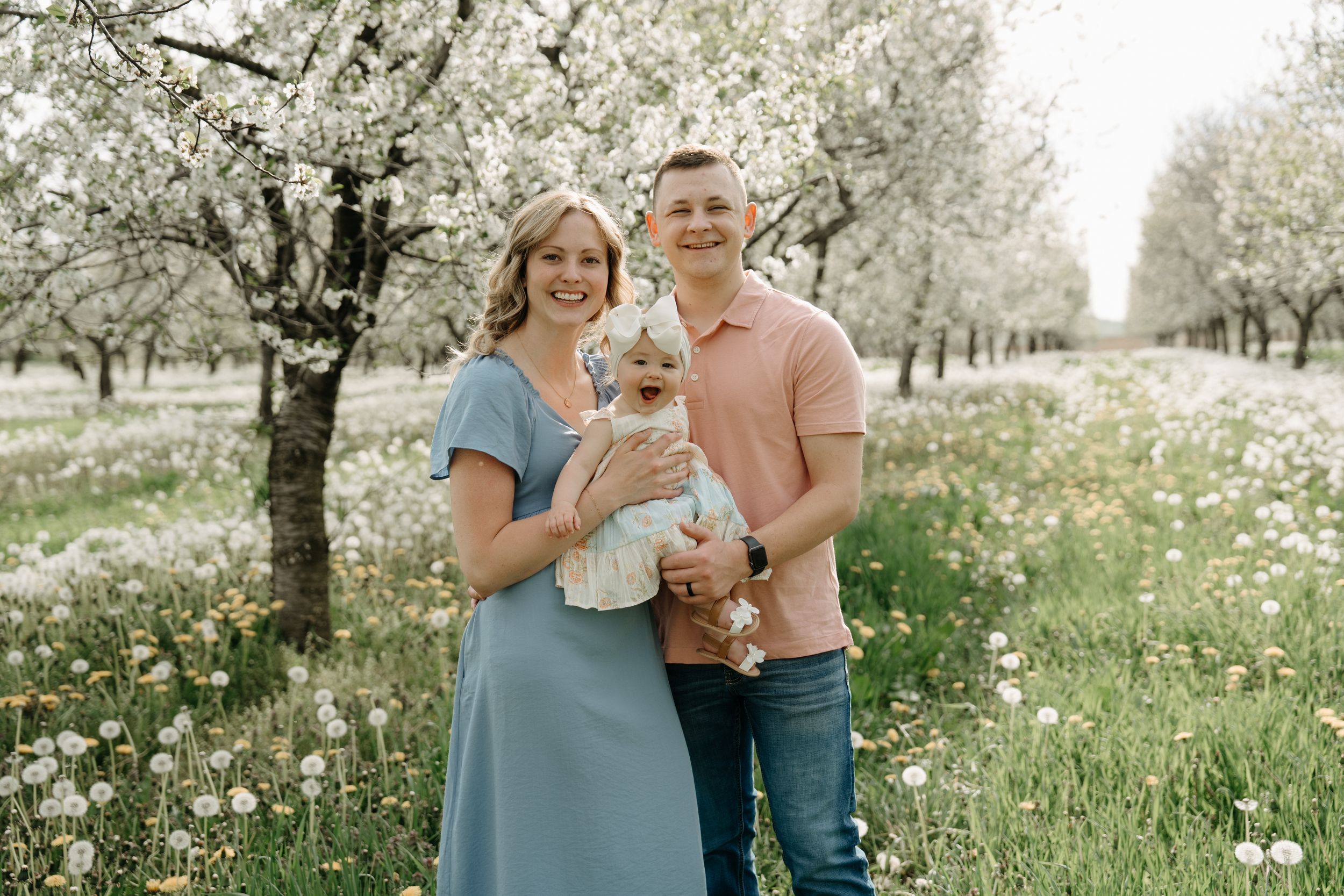 A couple in casual clothing holding a baby while standing in an orchard with blooming cherry trees and wildflowers.