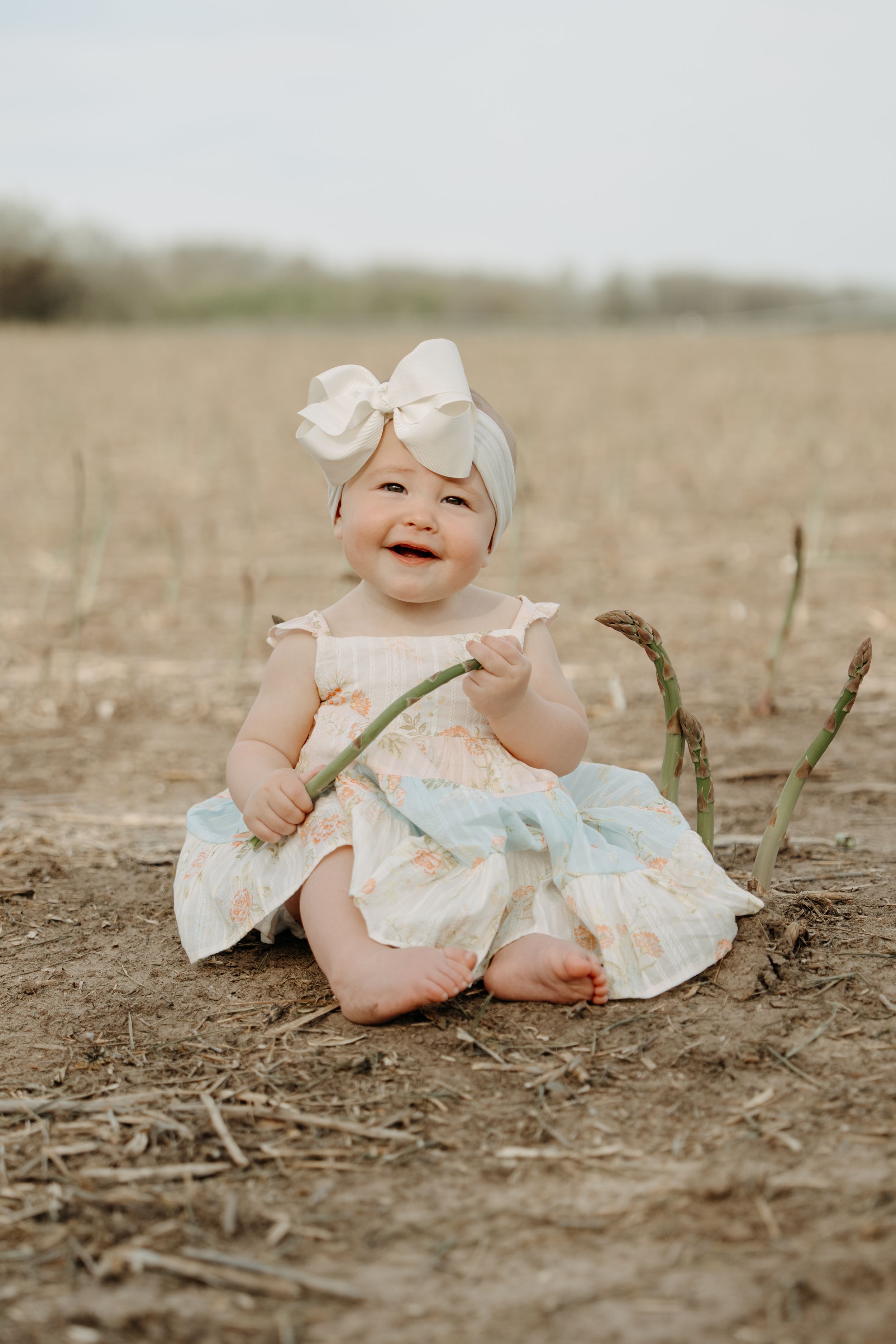 Baby wearing white dress and bow sits smiling in field while holding a small branch in their hands.