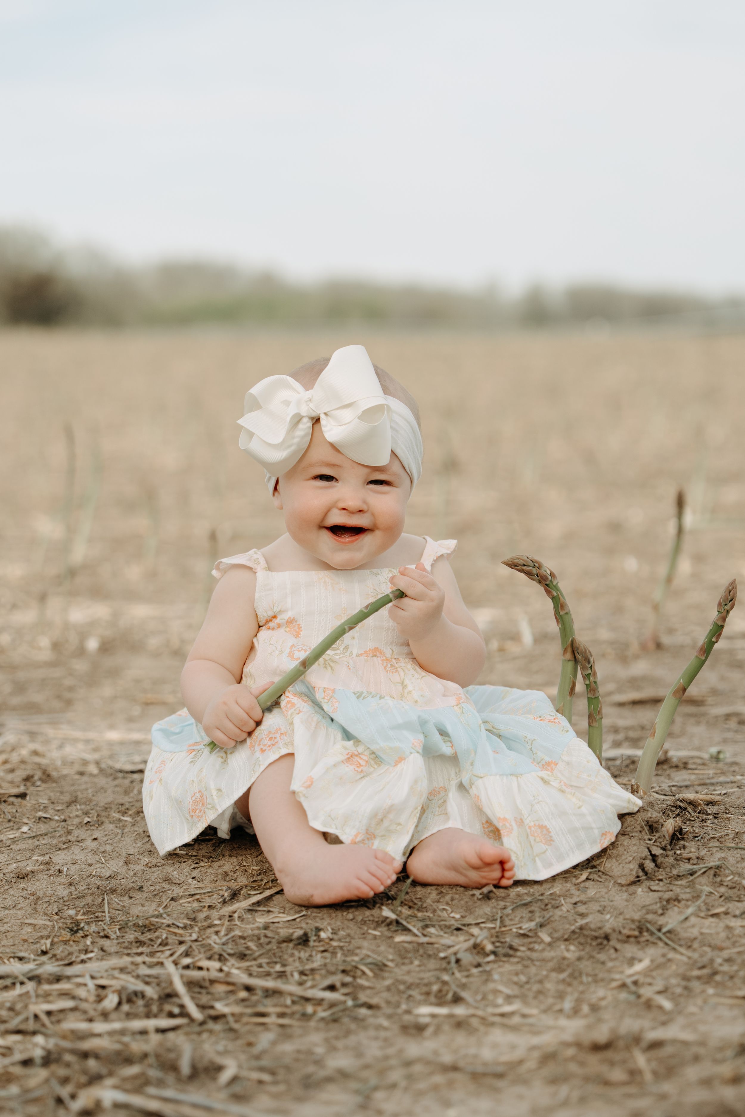 A baby wearing a white bow headband and floral dress sits happily in a rustic outdoor field setting.