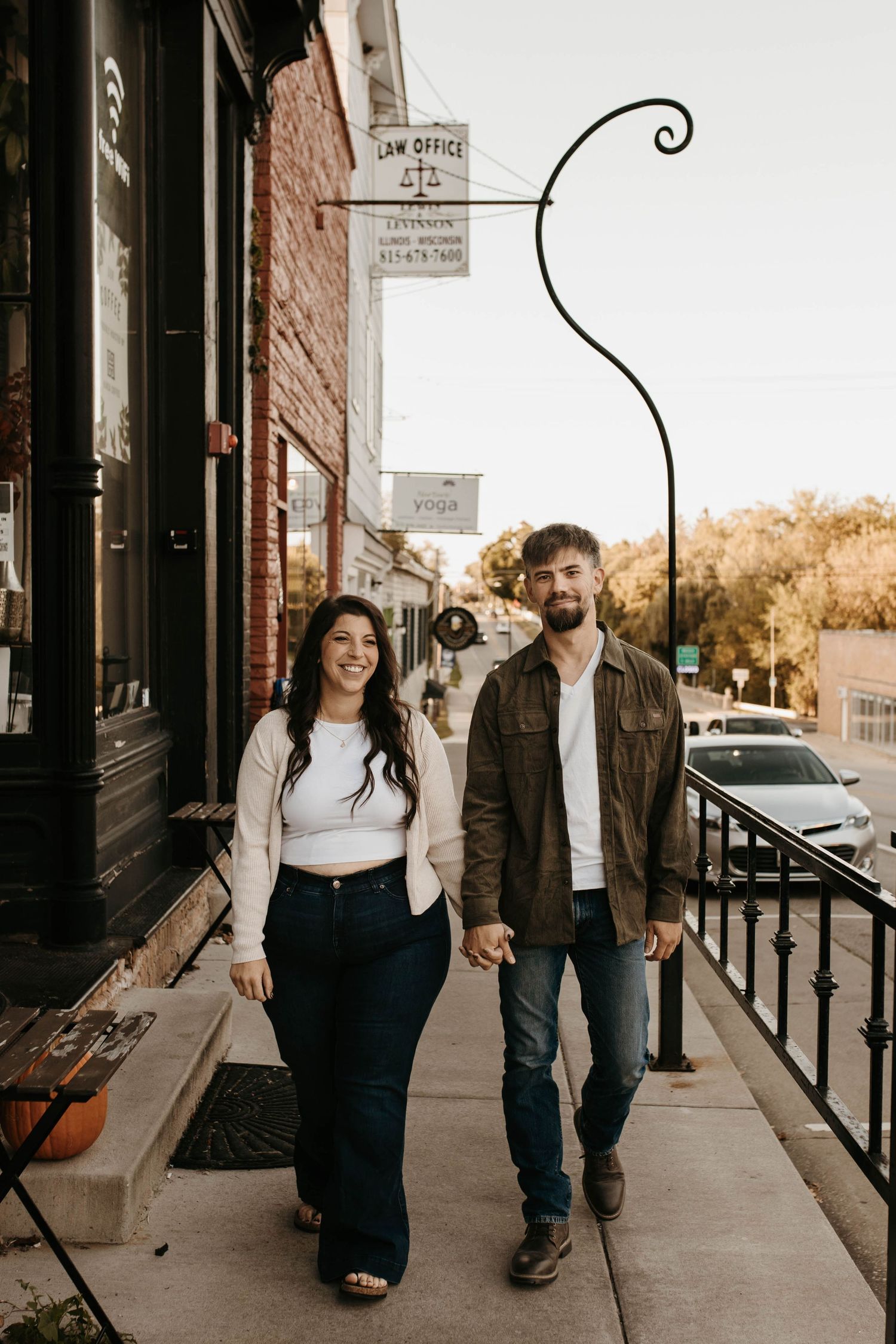 Couple holding hands and smiling while walking along a charming small-town sidewalk in autumn.