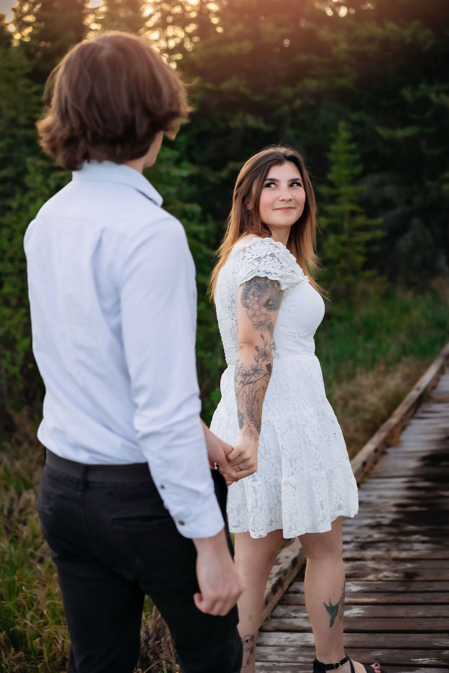 Couple in wedding attire holds hands on wooden boardwalk at sunset with forest backdrop and visible tattoos.