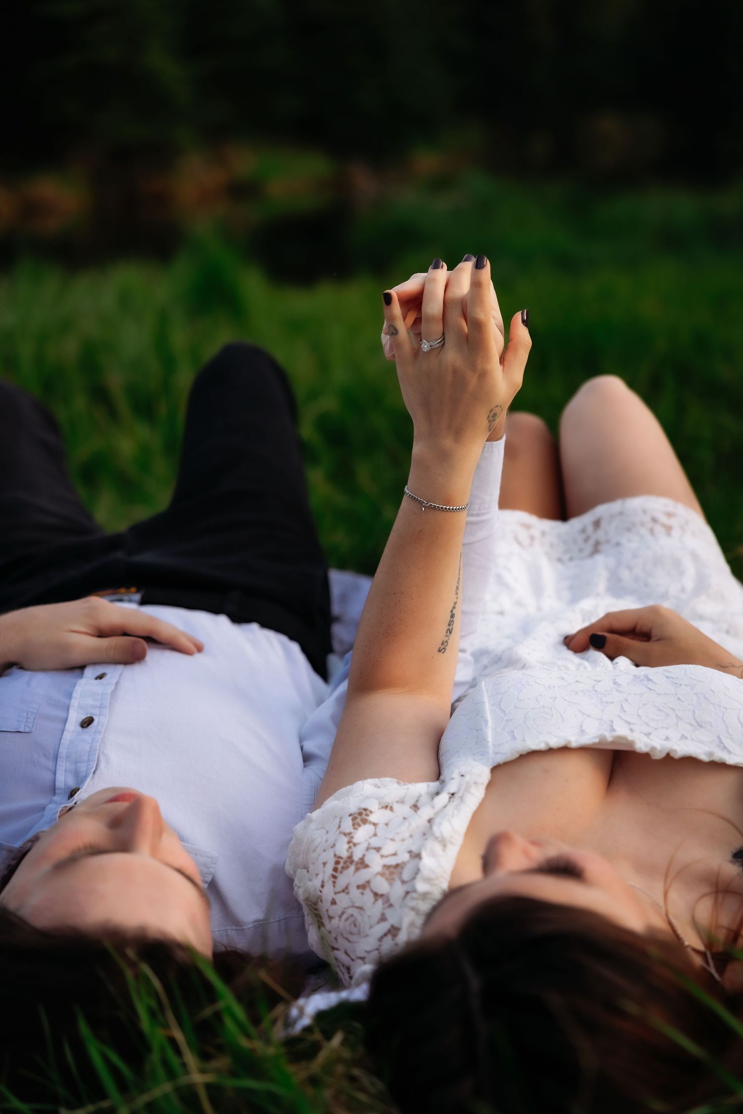 A romantic couple lying in grass holding hands, wearing white and denim, photographed from above.