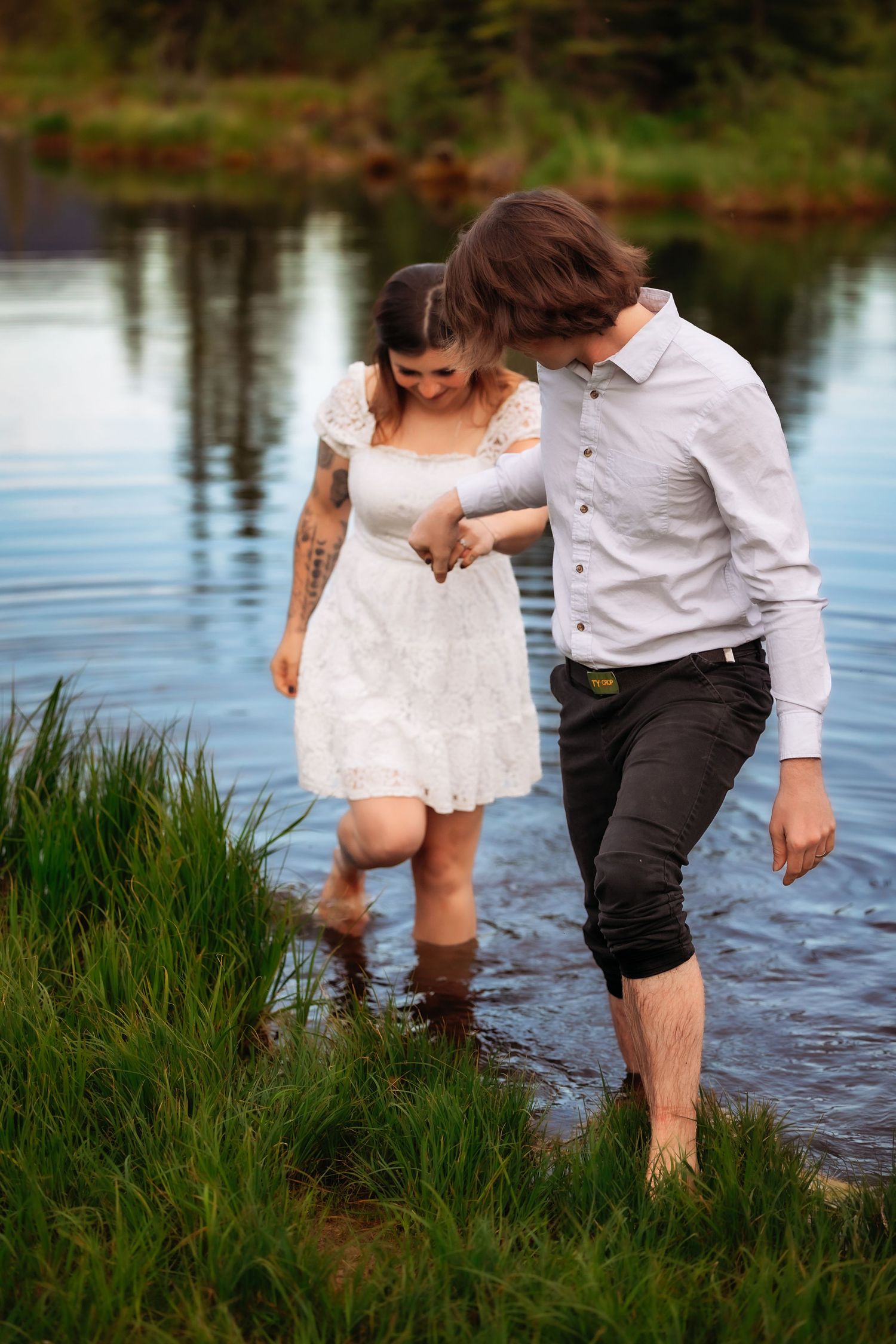 A couple wades together in shallow lake water, the woman in a white dress and man in rolled-up dark pants.