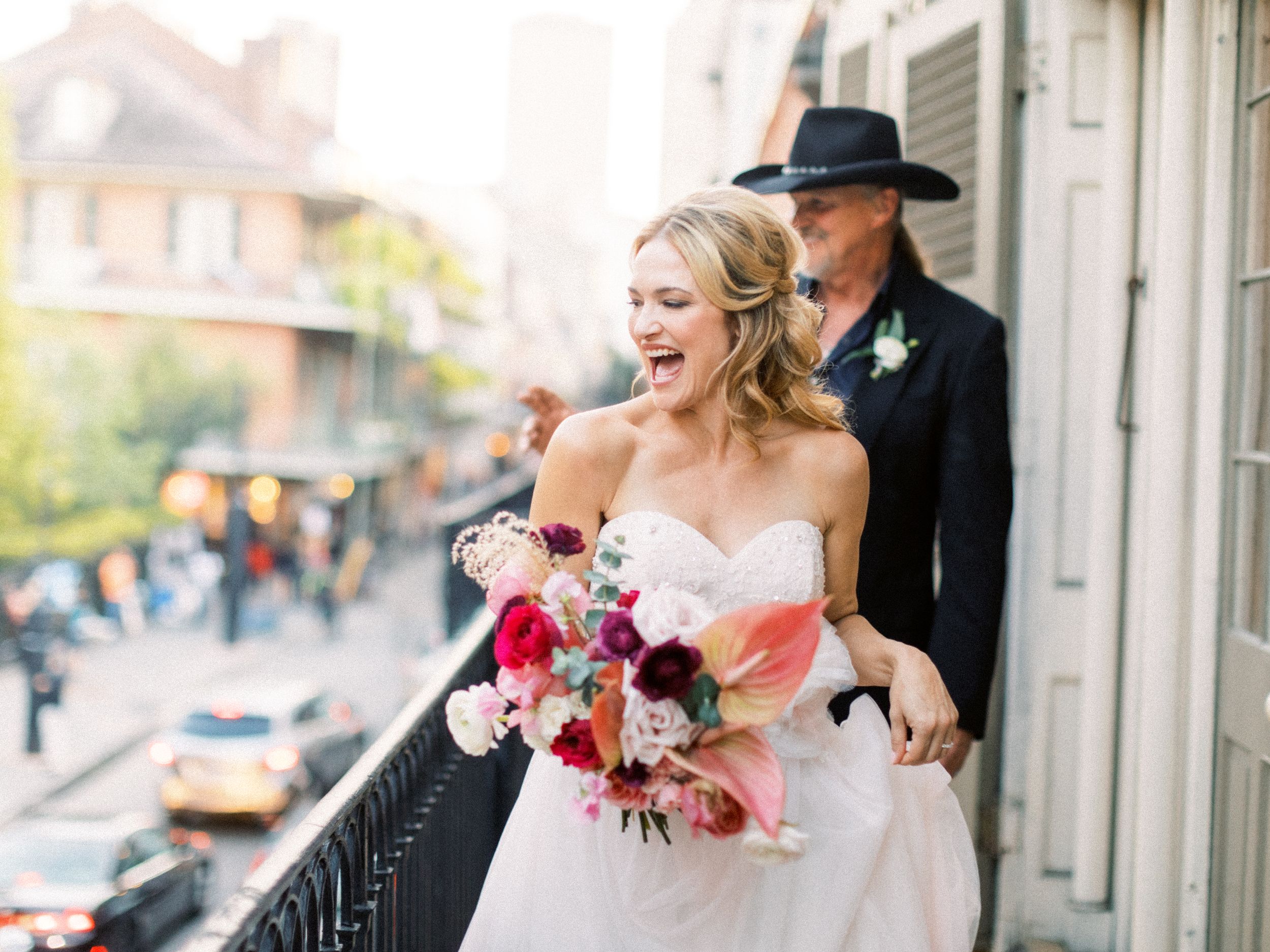 A bride in a white wedding dress and a groom wearing a dark Cowboy hat share a joyful moment on a balcony overlooking the French Quarter