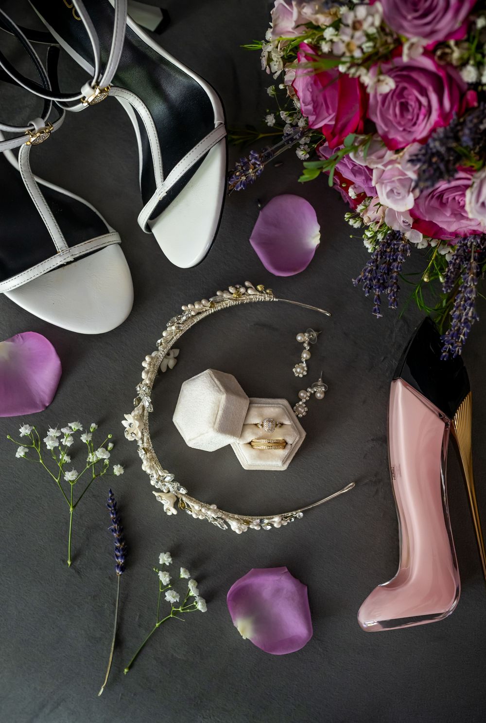 Wedding styling flat lay with purple roses, bridal shoes, jeweled headpiece and accessories on dark background.