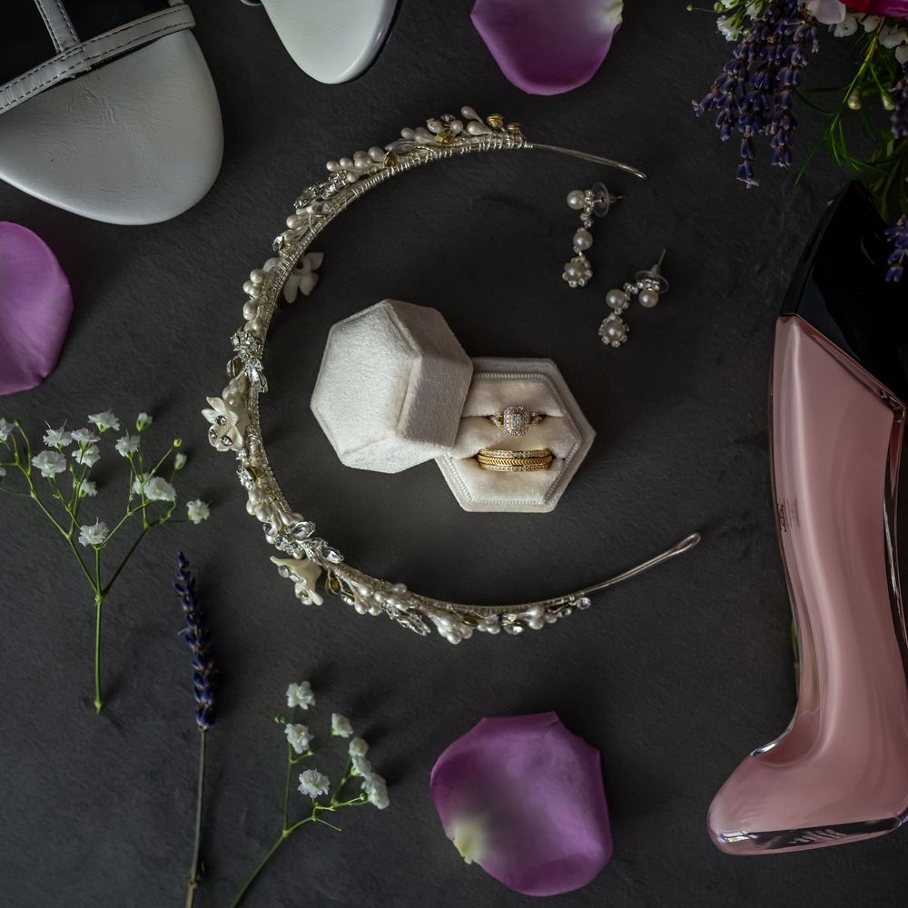 Wedding accessories including a crystal headpiece, ring box, earrings, and purple flower petals arranged on dark surface.