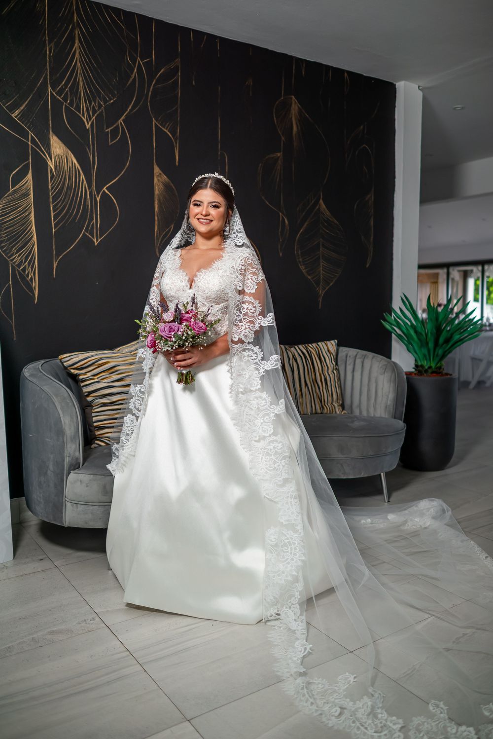 Elegant wedding dress with lace details and long train against black and gold wall with modern seating.