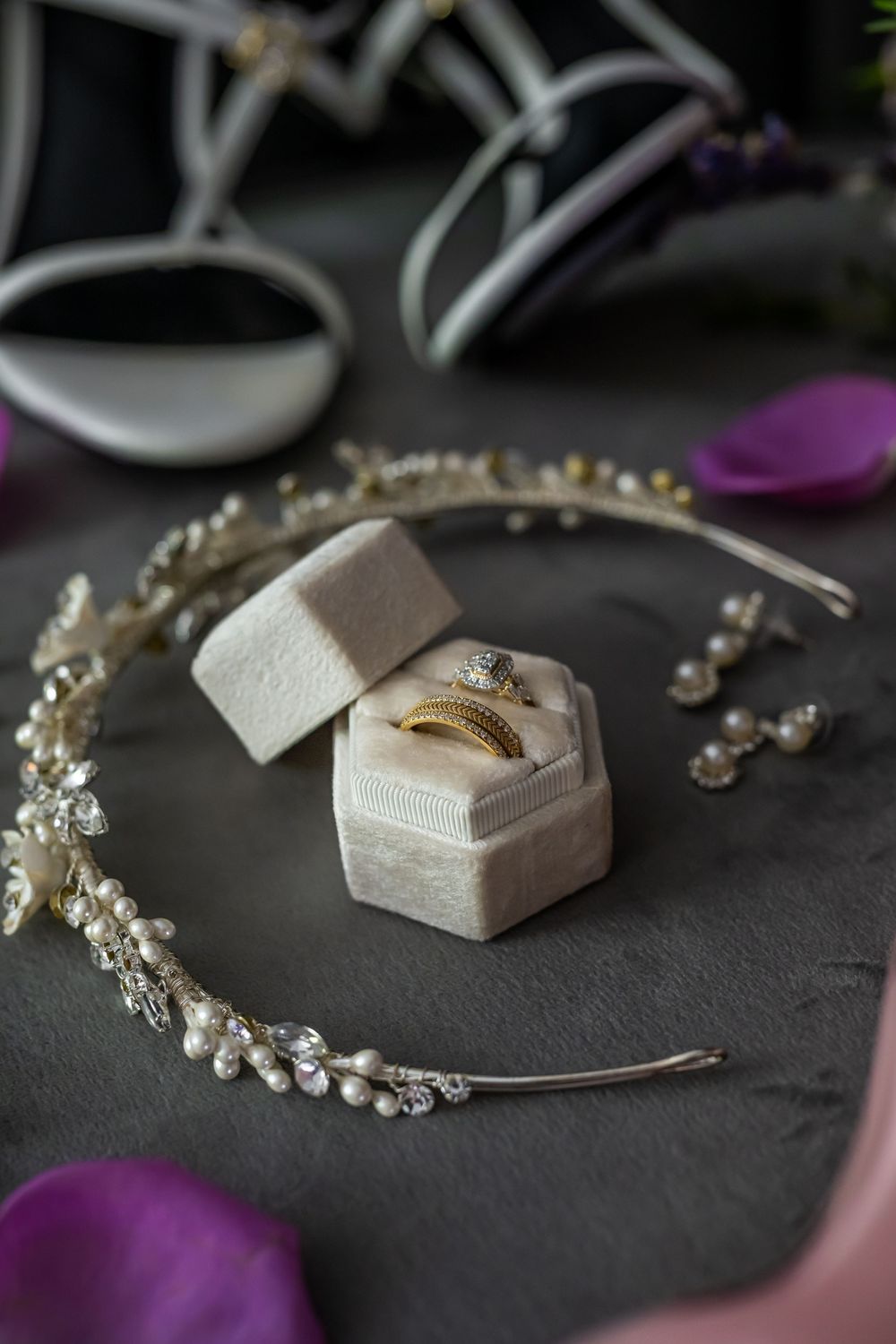 Wedding rings displayed in a velvet hexagonal box with crystal bridal headpiece and purple petals on dark surface.