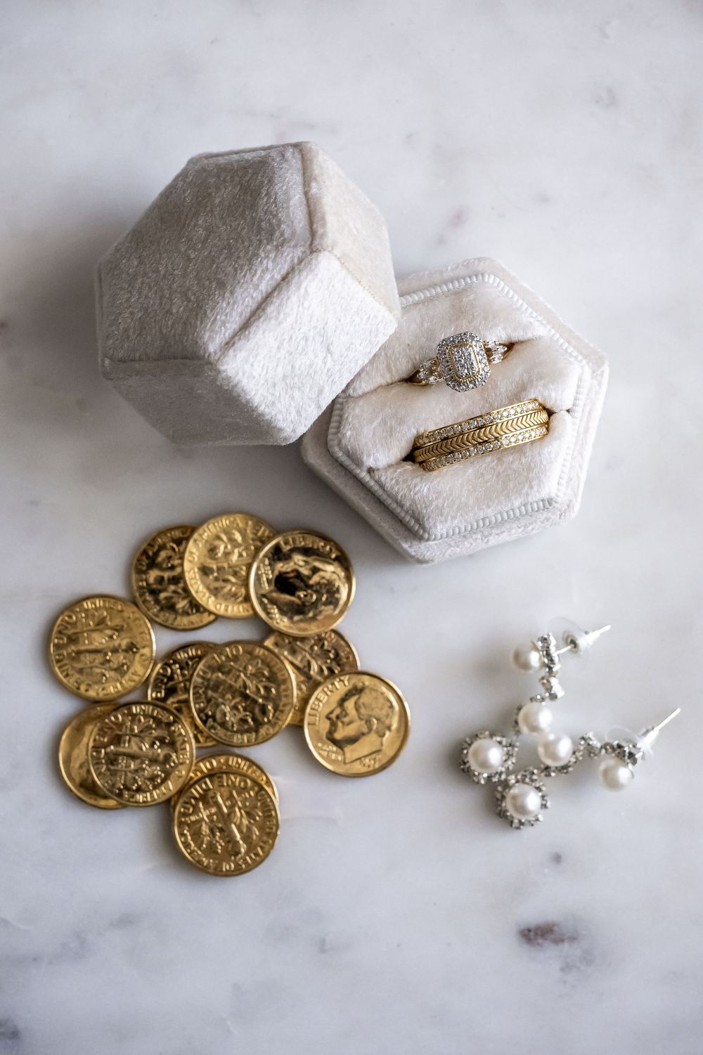 Wedding accessories including gold coins, pearl earrings and a hexagonal ring box on marble surface.