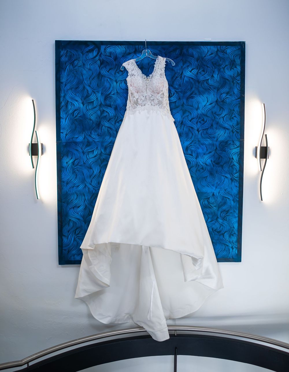 Elegant white wedding dress with lace bodice displayed against blue floral wallpaper with modern wall sconces.