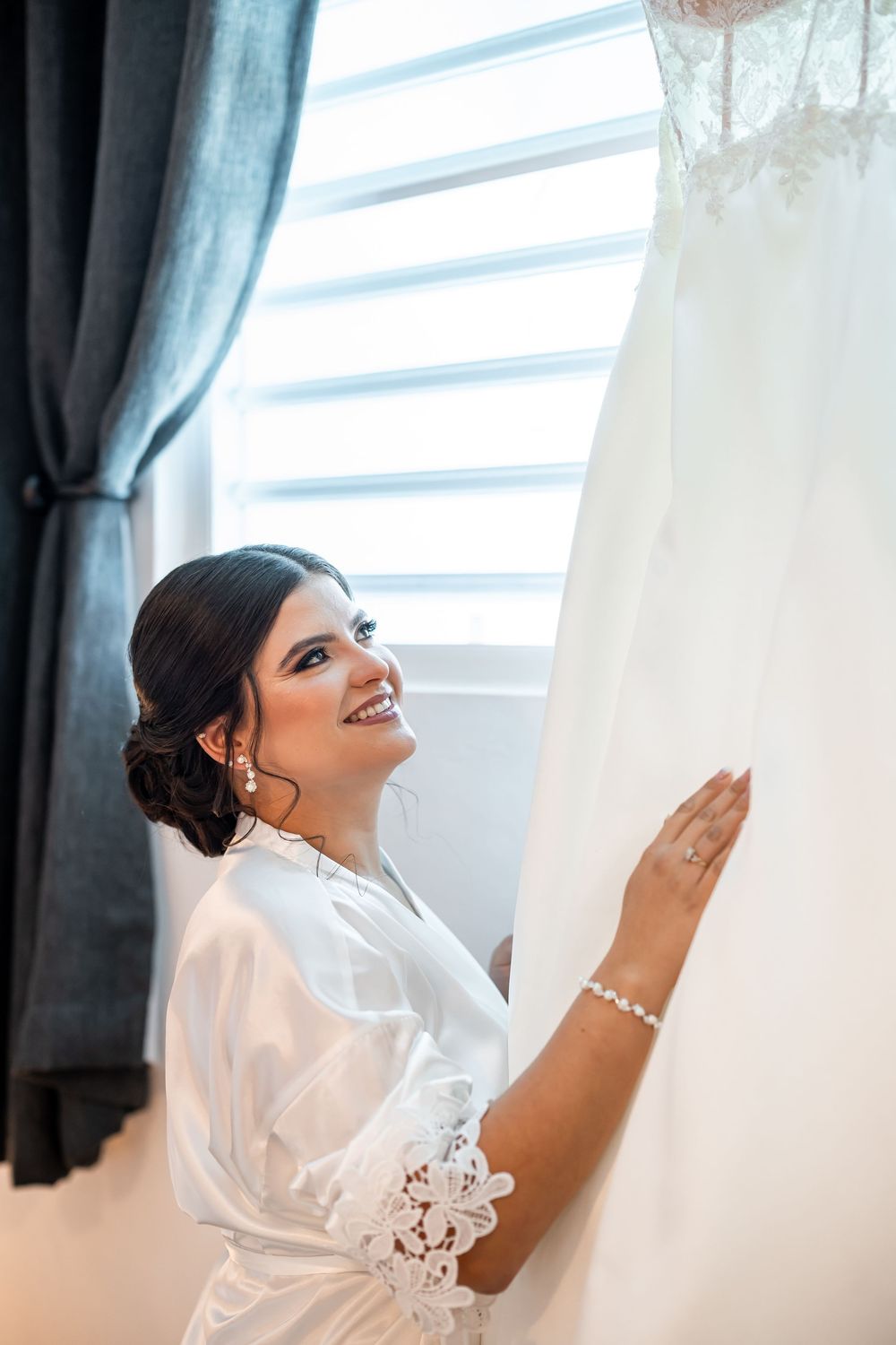 White silk wedding dress hanging by window with blue curtains as morning light streams in.