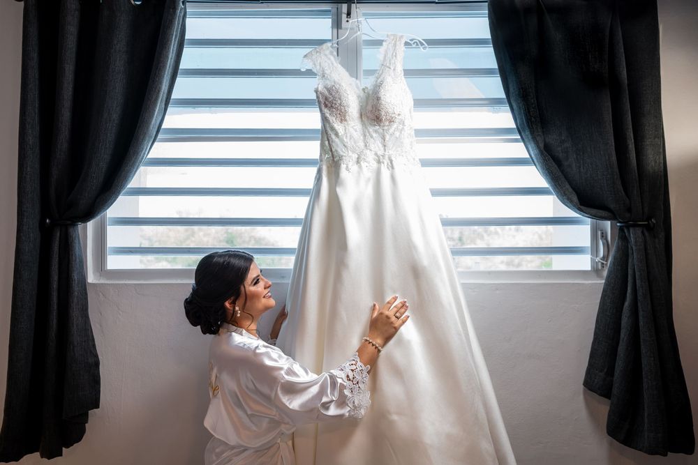 White wedding gown with lace details hanging from window between dark curtains.