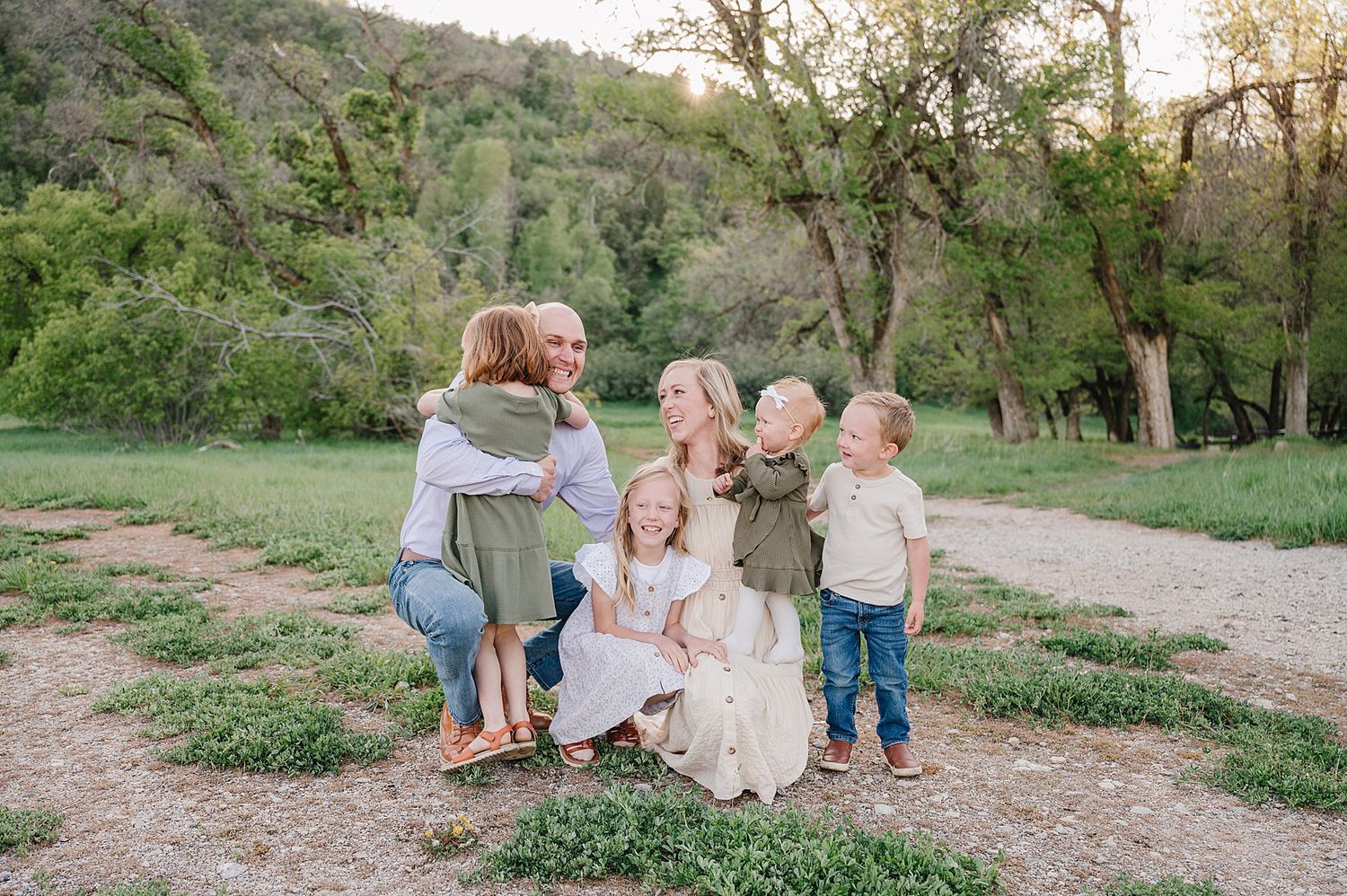 A family shares a sweet embrace on a dirt path surrounded by lush green hills and trees at sunset.