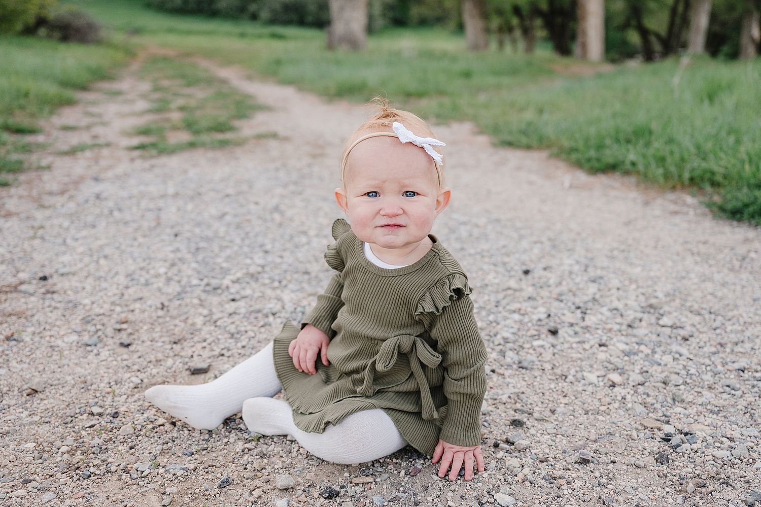 A small child in an olive green dress sits on a gravel path surrounded by green trees and grass.