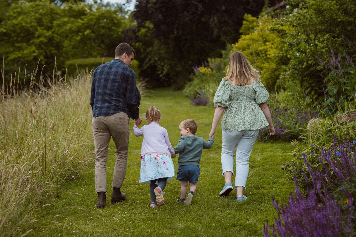 A family walks together down a garden path bordered by purple flowers and tall grasses on a summer day.