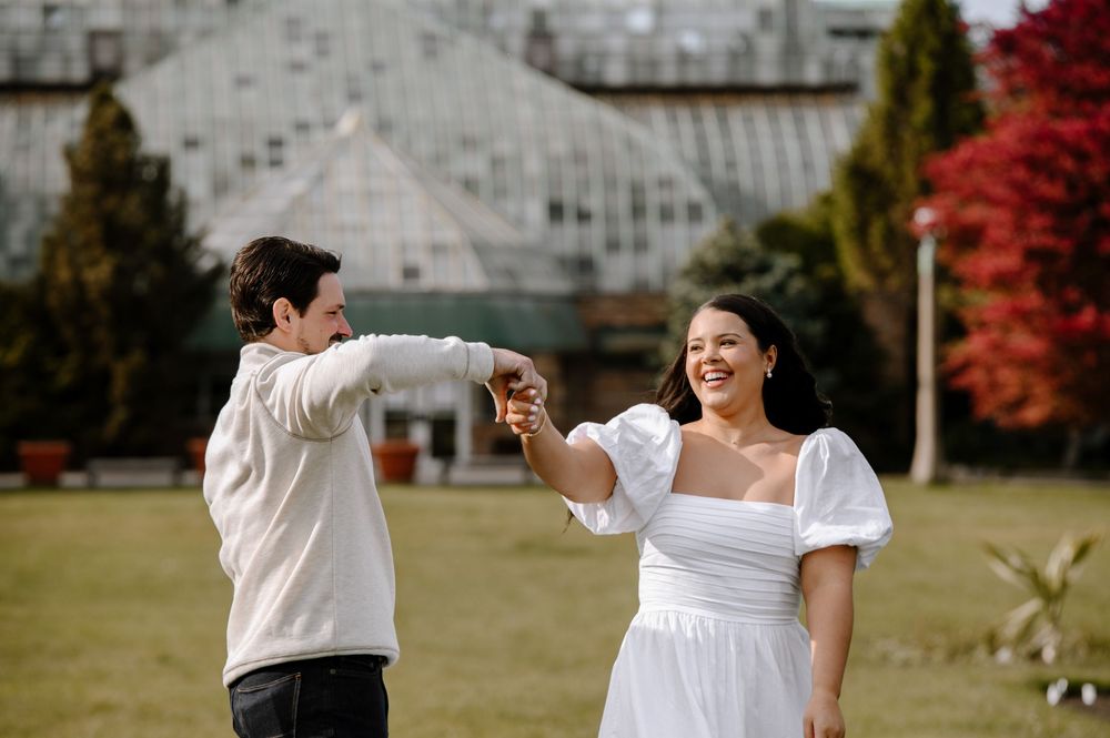 A couple holds hands and dances joyfully on a lawn in front of a glass conservatory building.