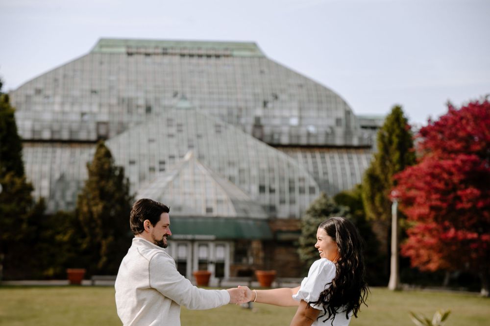 A couple stands on lawn facing each other with a large glass building and autumn trees in background.