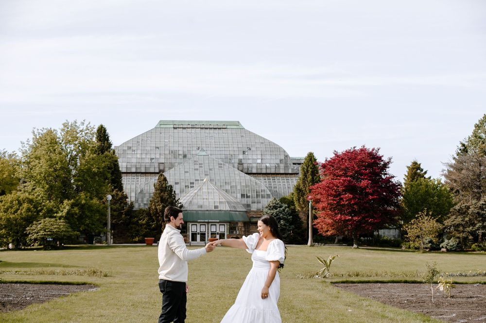 A couple poses in front of a large greenhouse with colorful autumn trees.