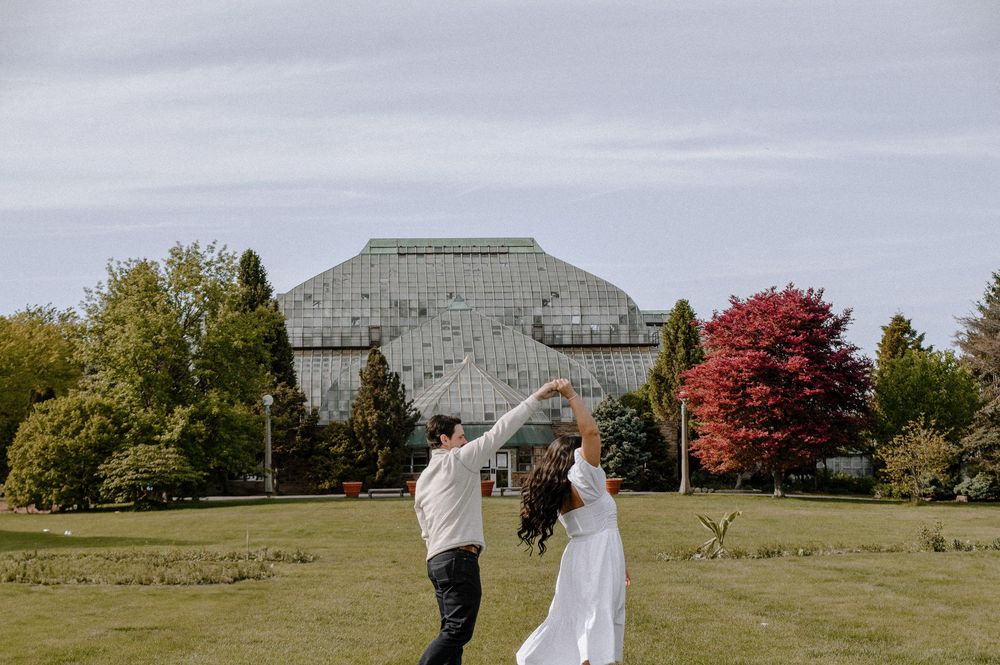 A couple dances playfully on a grassy lawn in front of a large glass building on an overcast day.