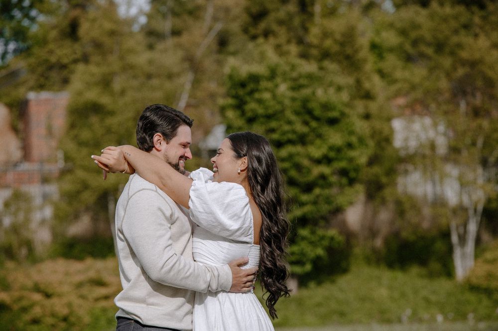 A romantic couple embrace and laugh together outdoors with green foliage in the background.