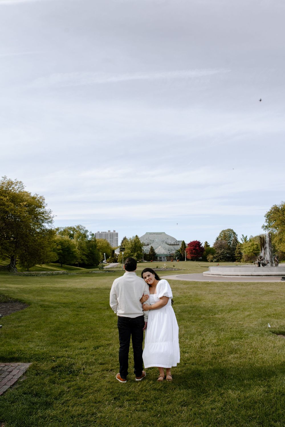 Two figures walk together across open grassy field with buildings in background.