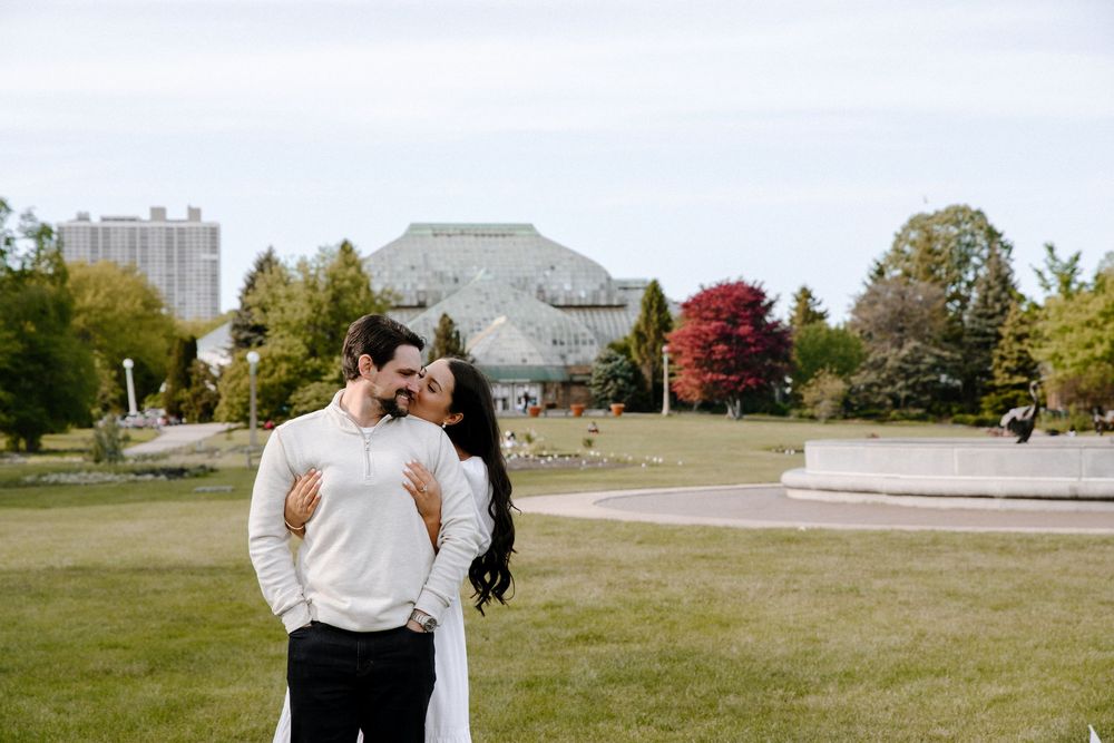 A couple shares a romantic moment in an open grassy park with fall foliage and buildings in the background.