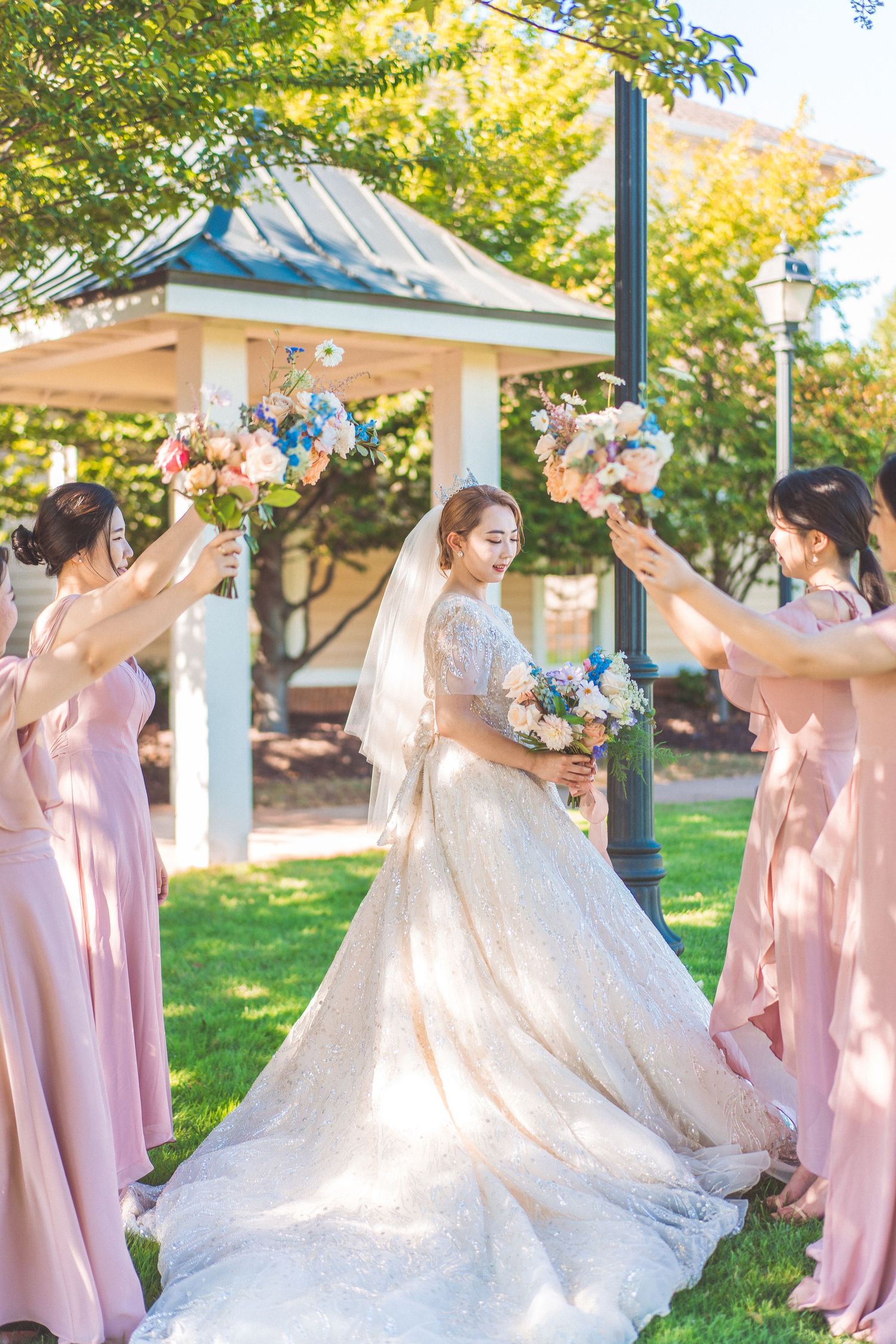 Bride in flowing white gown with bridesmaids in pink dresses holding bouquets aloft.
