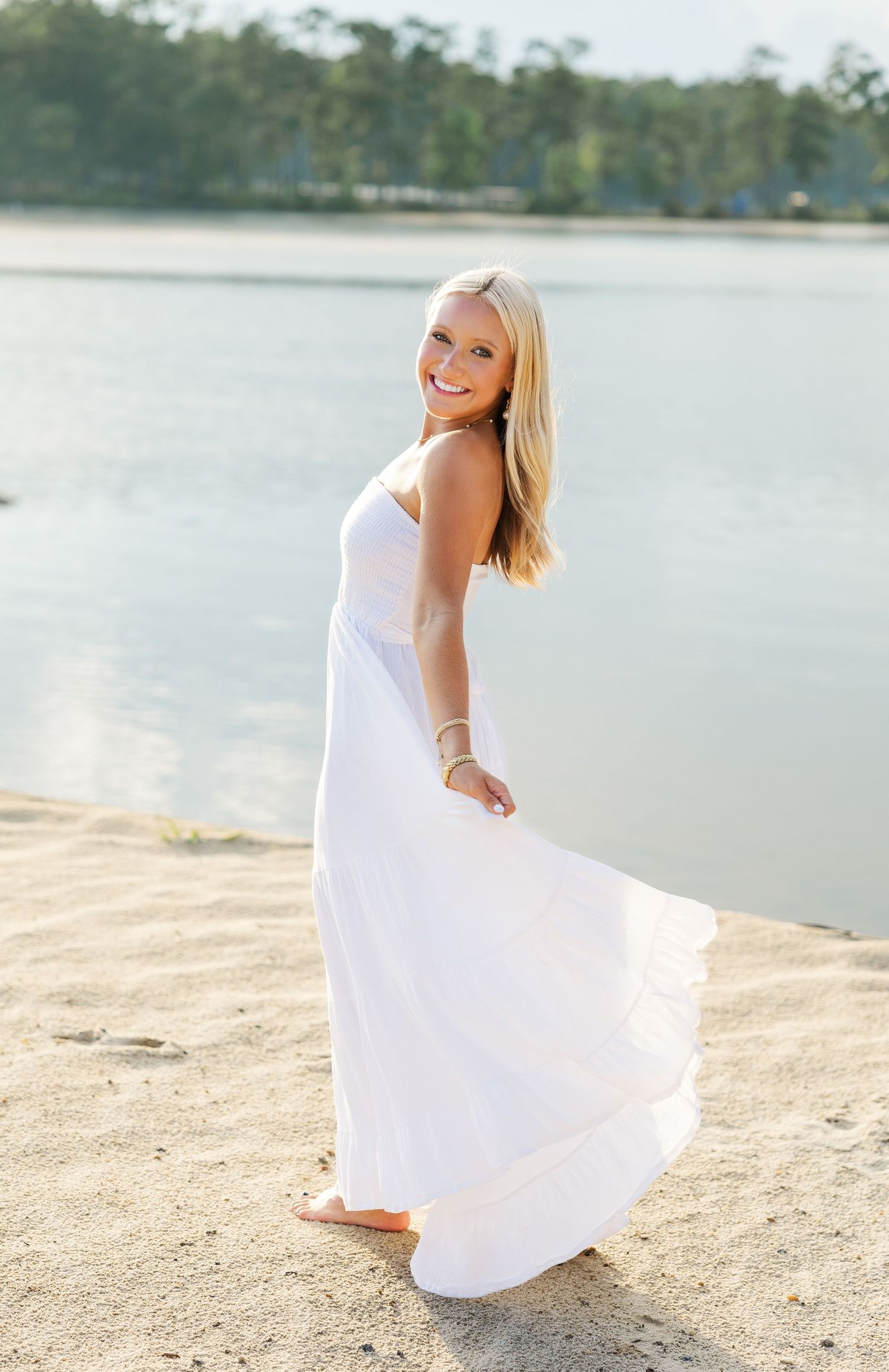Woman in flowing white dress poses gracefully on beach shore with lake and trees in background.