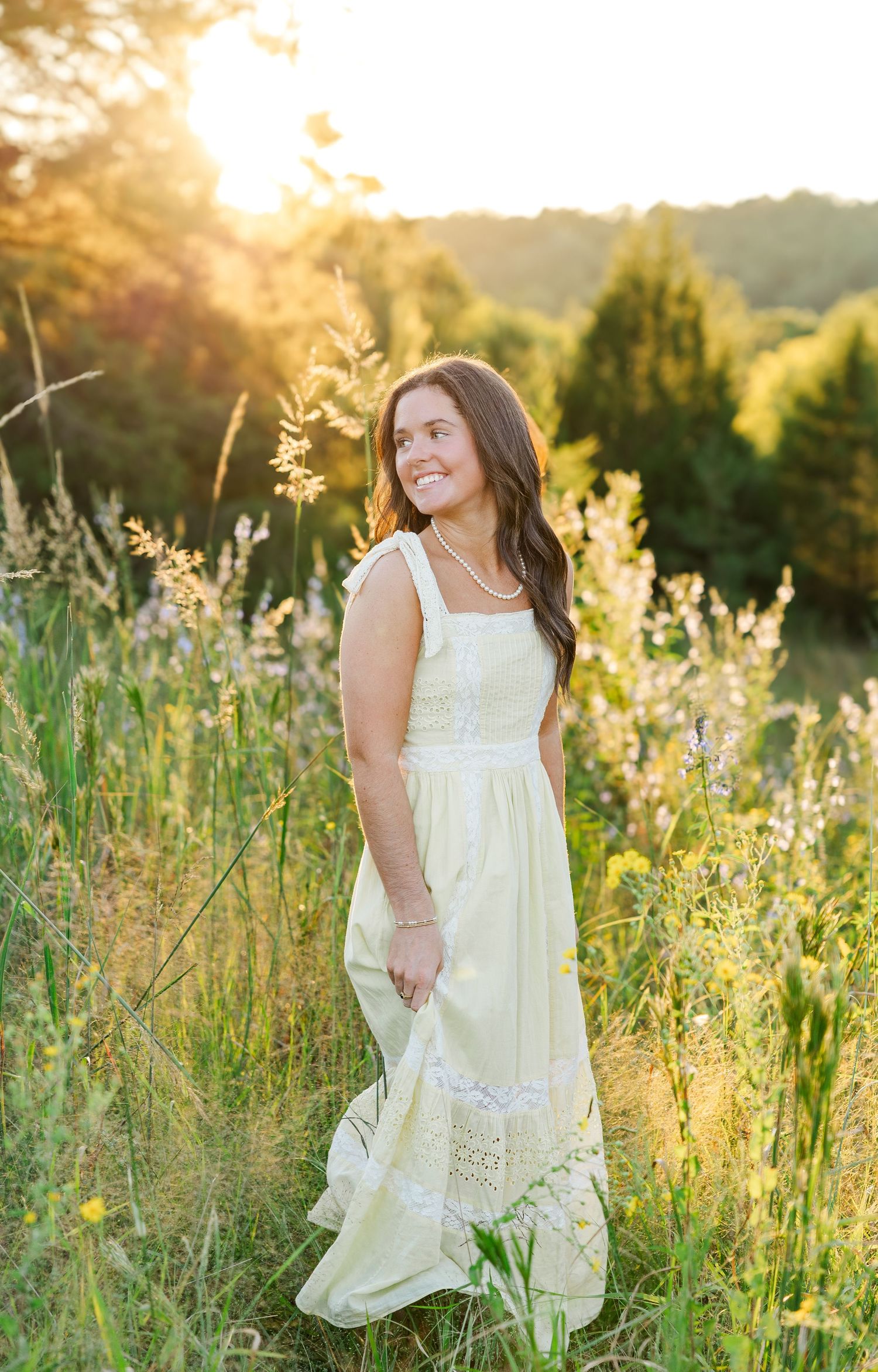 A figure in a flowing white dress stands in a golden sunlit meadow with tall grass and wildflowers at sunset.