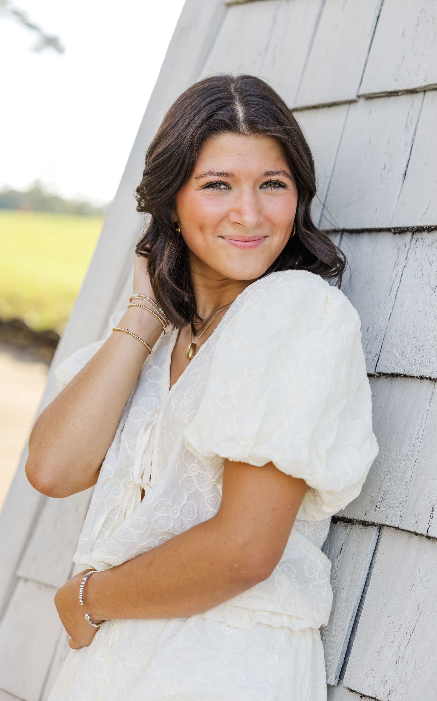 Portrait of a young woman in a white puff-sleeve dress leaning against a gray shingled wall on a sunny day.