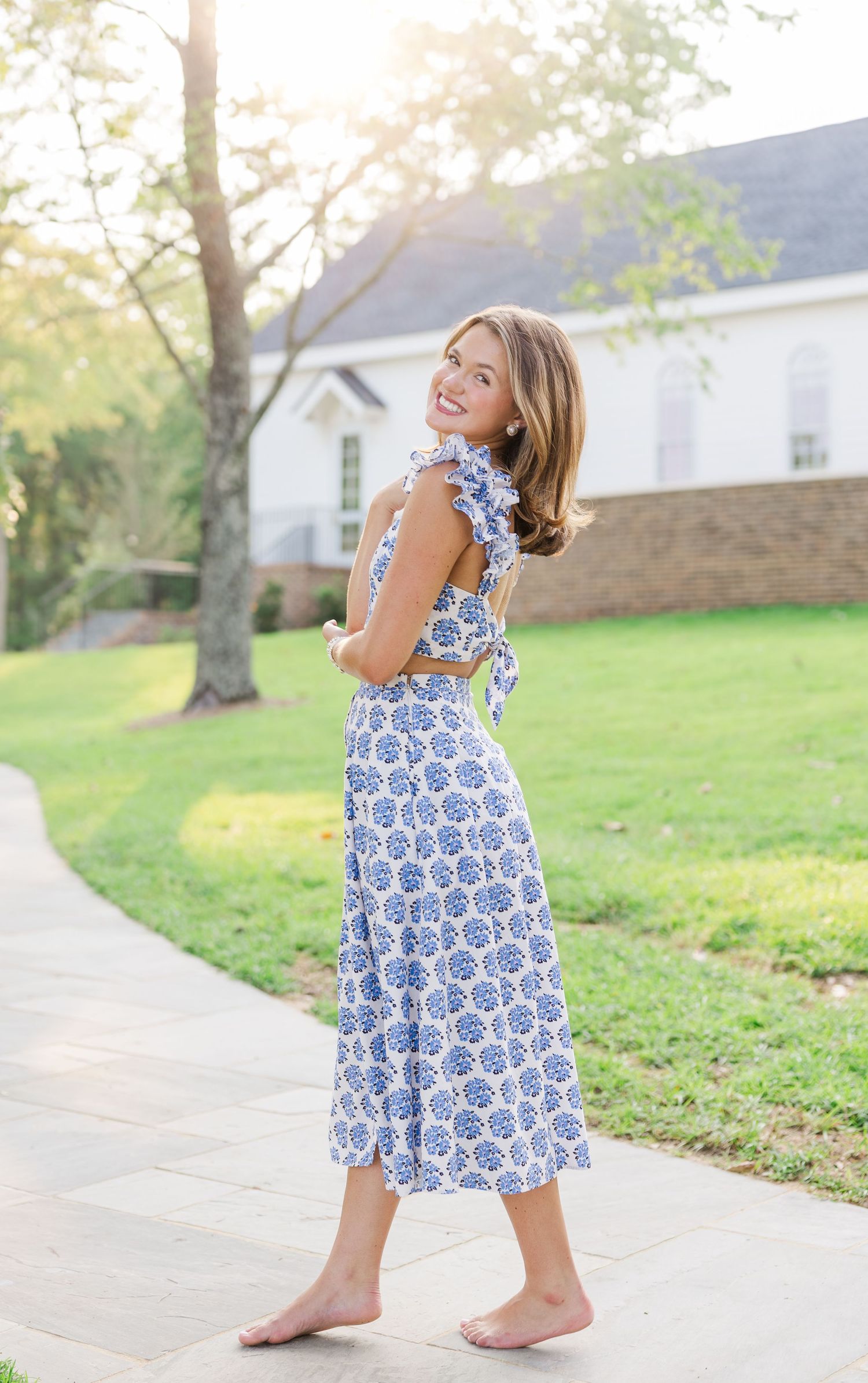 A woman in a blue and white floral dress poses barefoot on a garden path during golden hour lighting.