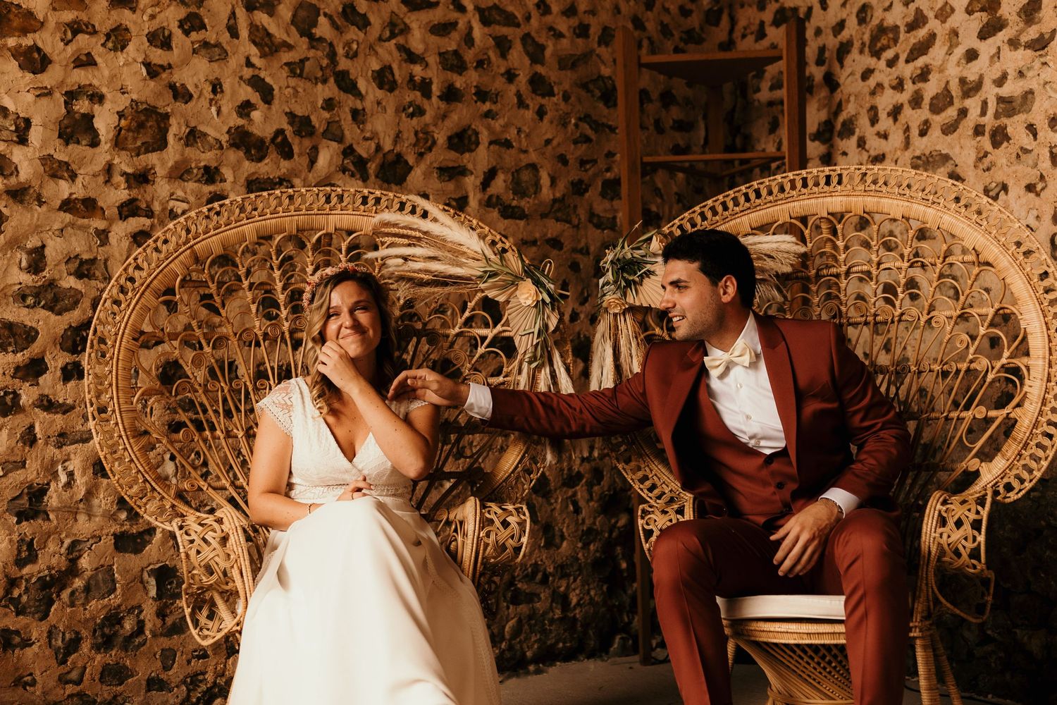 Wedding couple poses on rattan peacock chairs against stone wall interior.