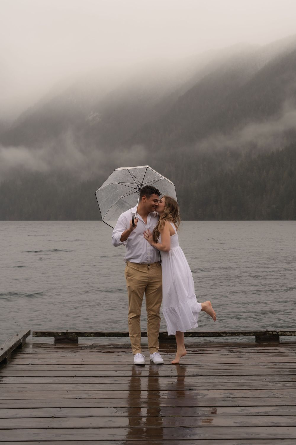 A romantic couple shares an umbrella on a misty wooden dock by a lake surrounded by foggy mountains.