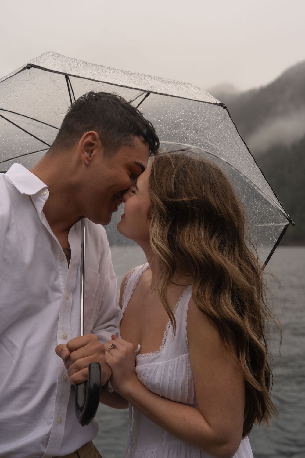 Romantic couple sharing an intimate moment under a clear umbrella by a misty lake.