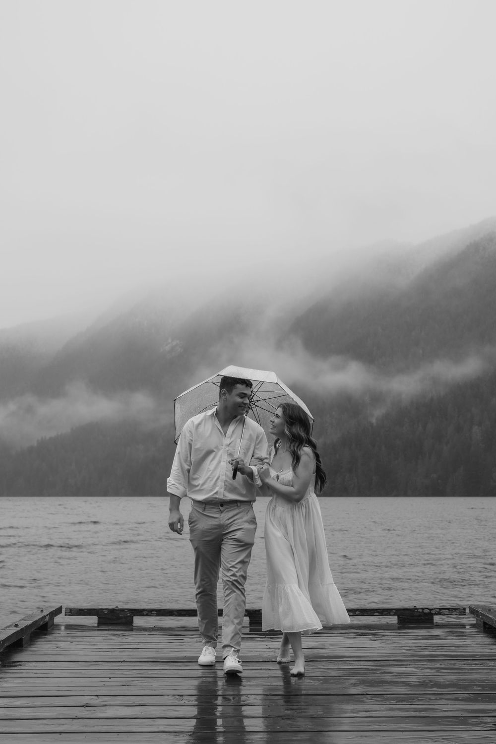 Romantic couple shares umbrella while walking on misty lakeside dock in black and white photography.