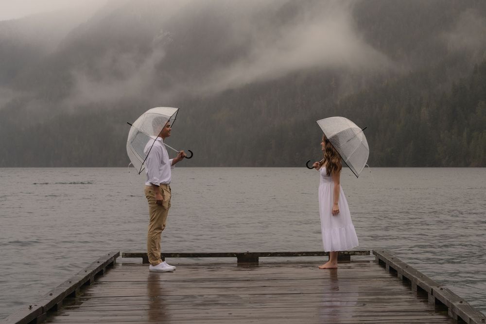 Two people with white umbrellas stand on a misty lakeside dock surrounded by mountains in moody weather.