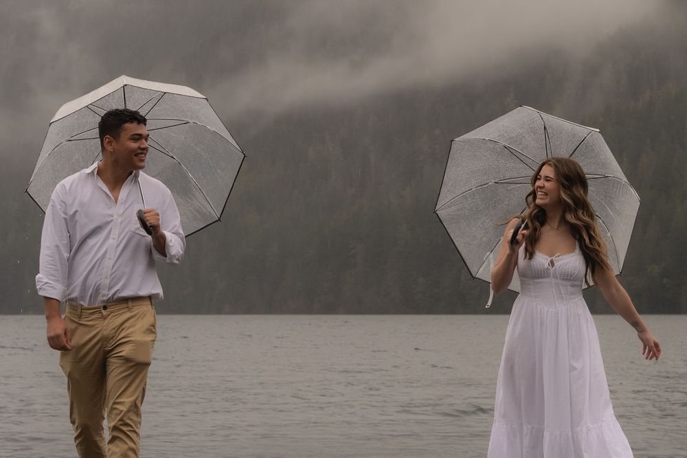 A couple stands with white umbrellas by the water against a moody gray sky.