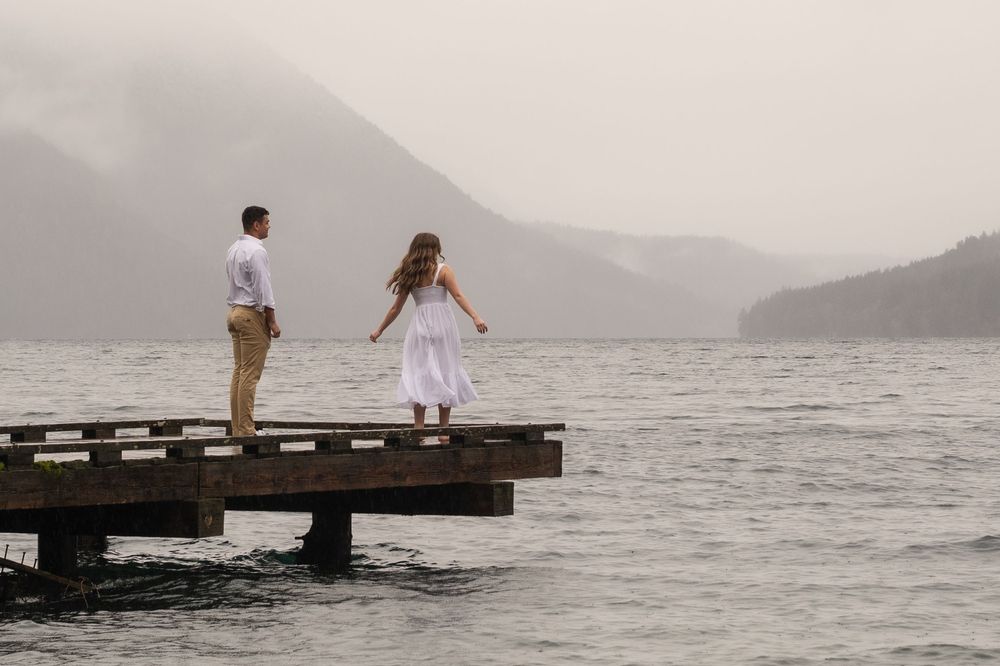 Two people stand together on a wooden pier overlooking a misty mountain lake on an overcast day.