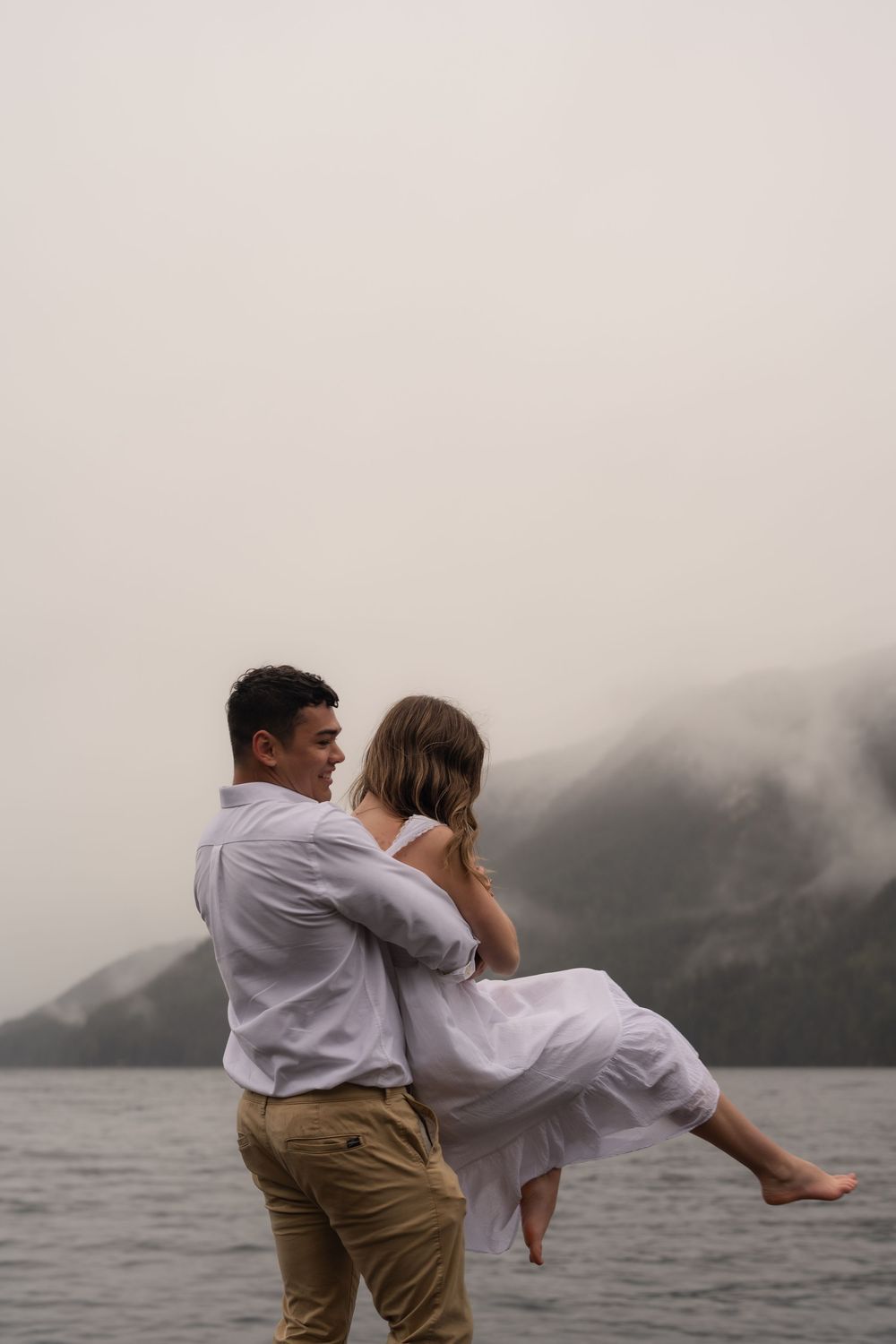 A couple in white clothing embraces playfully by a foggy lake while the man lifts his partner.