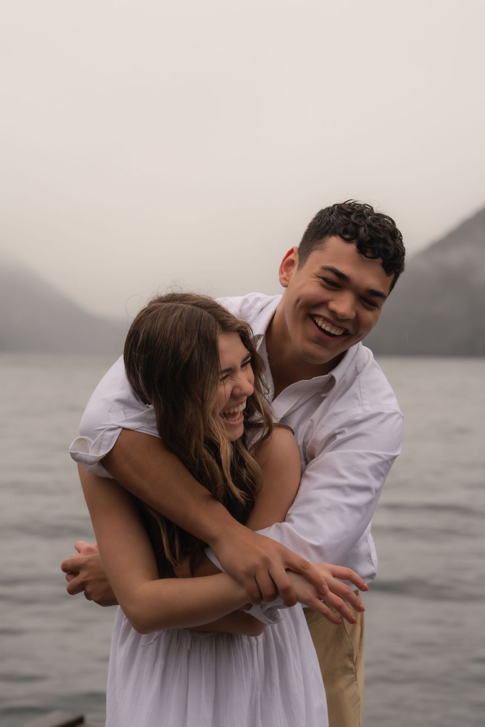 A romantic couple in casual white outfits share a joyful embrace by a misty lake with mountains in the background.