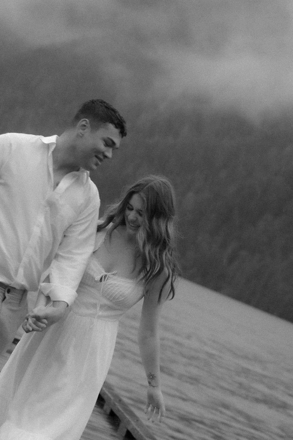 A romantic black and white photo of a couple in white clothing standing together by the water with mountains in the background.