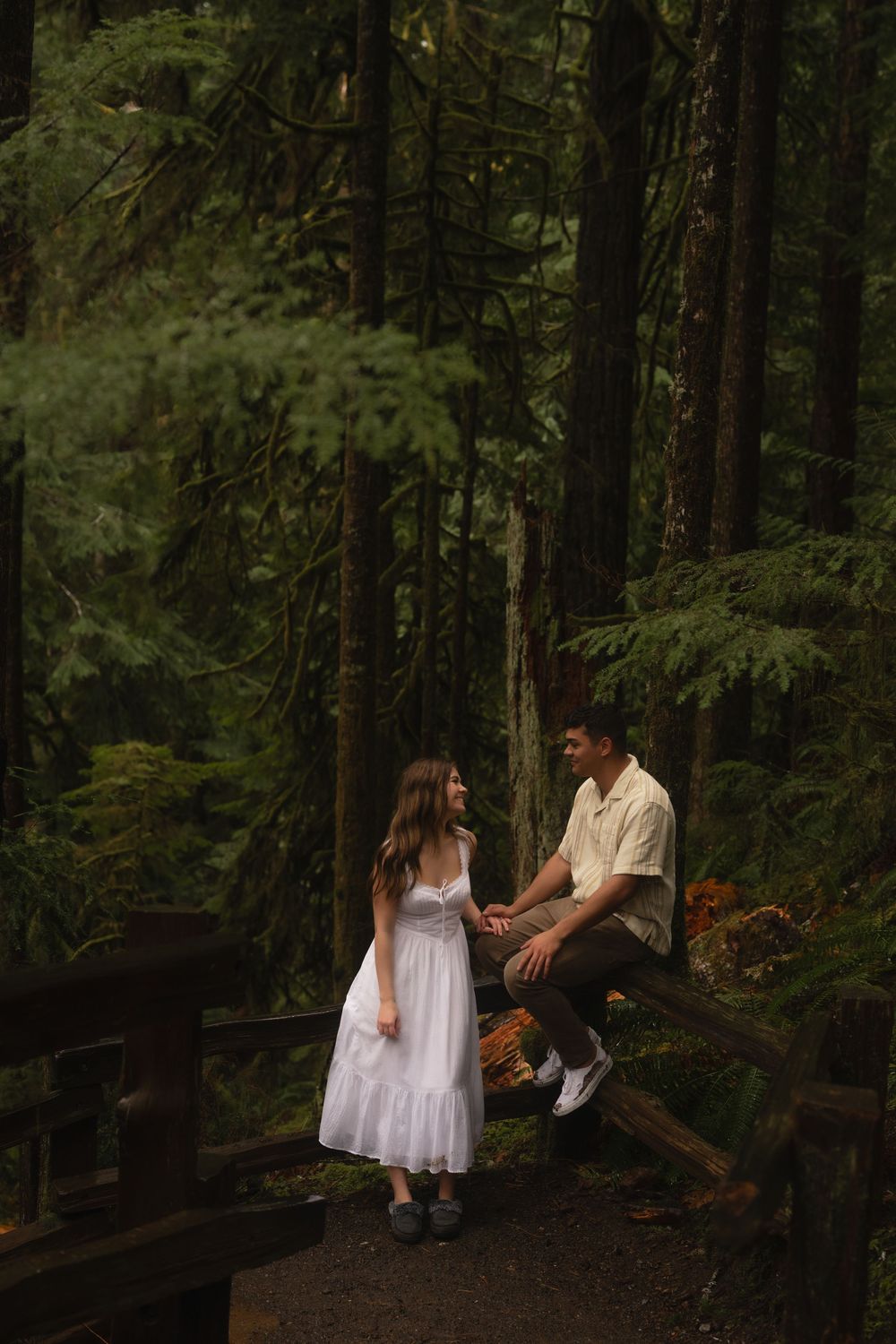 A romantic couple shares a tender moment while standing on a wooden bridge in a misty evergreen forest.