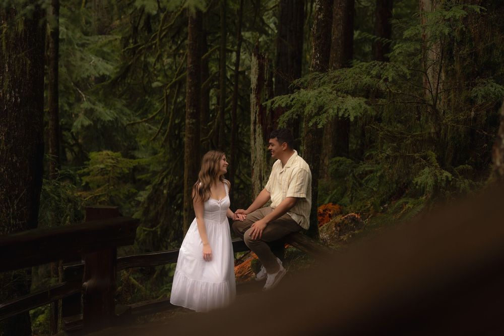 Two figures in formal attire share a romantic moment on a wooden bridge surrounded by lush forest greenery.