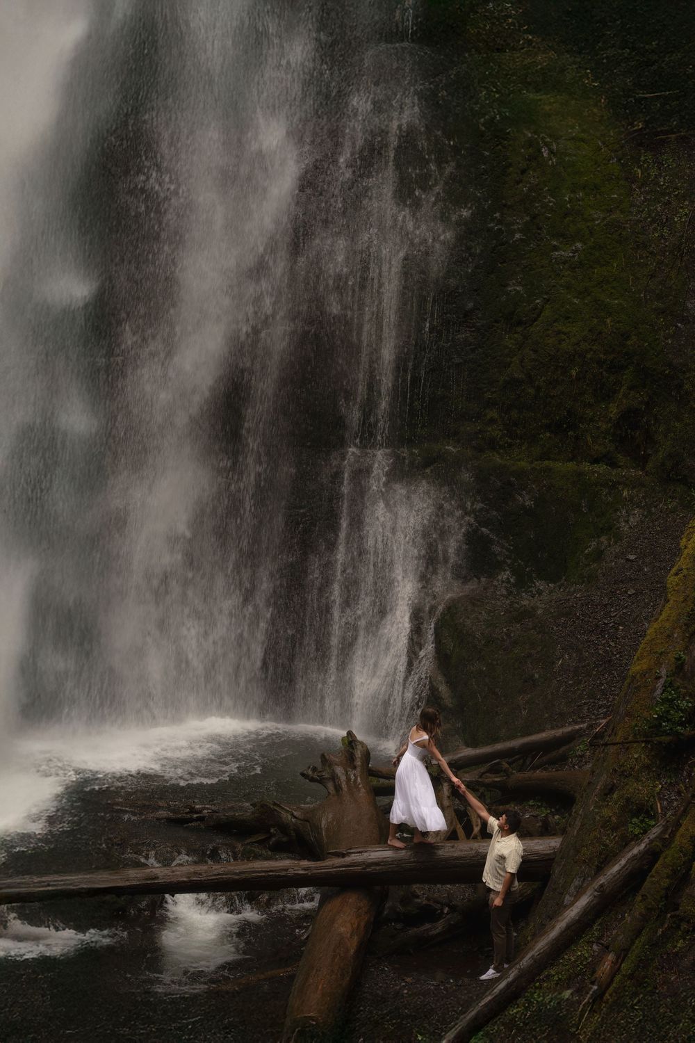A couple poses on fallen logs near a majestic waterfall in a scenic forest setting during a photo session.