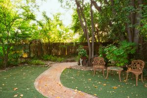 A curved brick pathway leads through a lush backyard garden with ornate metal chairs and a wooden privacy fence.
