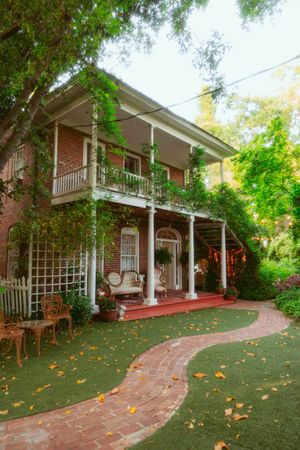 Scenic panorama of a classic brick colonial home with wrap-around porch and lush landscaping in a peaceful garden setting.
