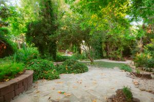 A winding stone pathway through a lush garden with raised flower beds and wooden benches beneath shady trees.